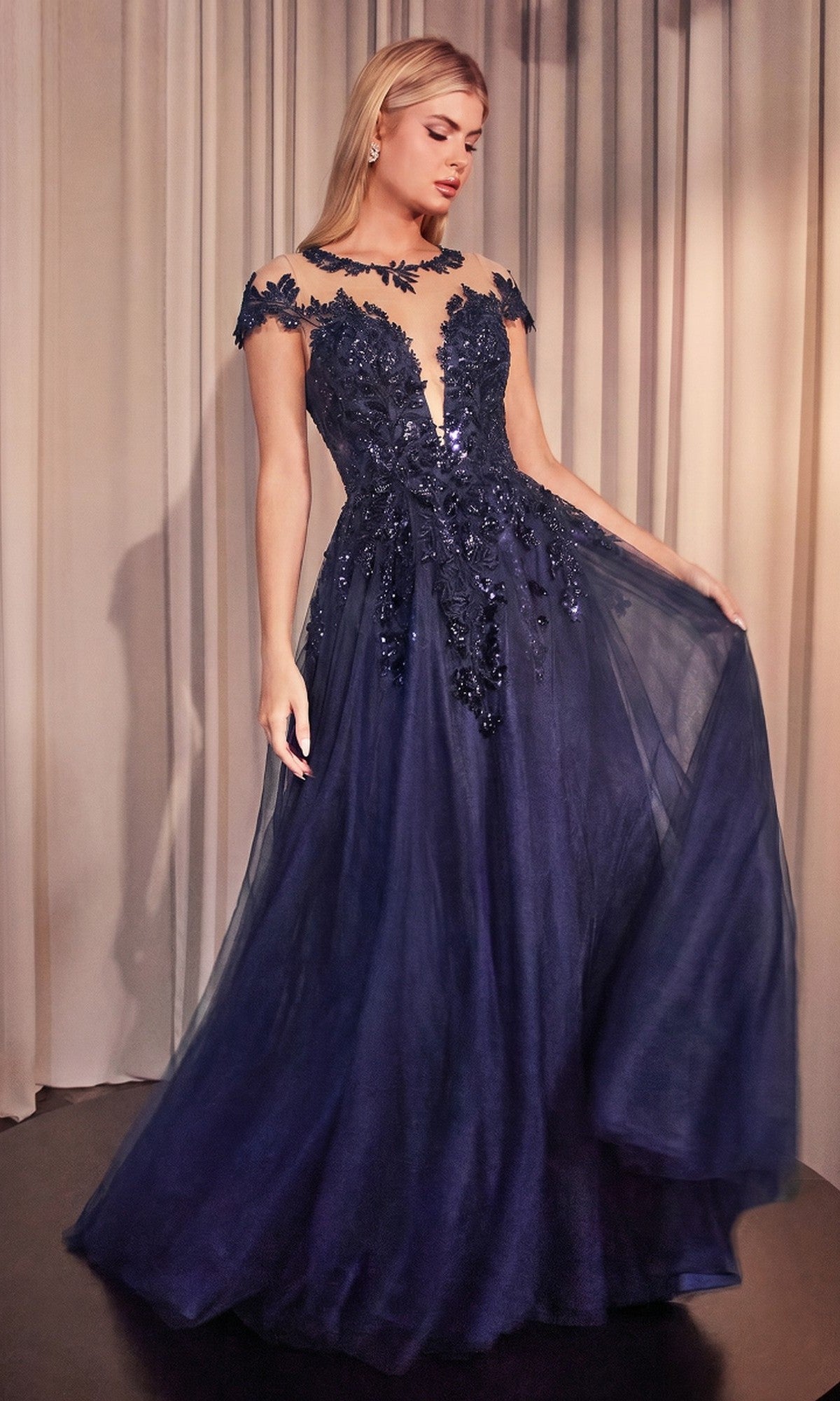 A woman stands in front of beige curtains wearing the Andrea and Leo A1475 navy long prom dress, featuring floral appliques, a sheer neckline, cap sleeves, and a flowing A-line skirt.